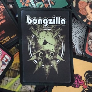 May include: Black and green embroidered patch with the text "bongzilla" in white letters. The patch features a green creature with large eyes and a skull-like head.