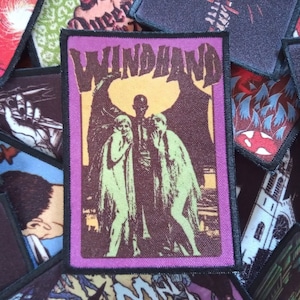 Peut inclure: Un patch brodé violet et marron avec le texte "WINDHAND" en jaune. Le patch présente une illustration en noir et blanc de trois personnages avec des ailes.