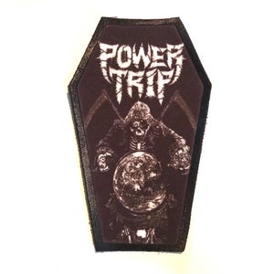 May include: A black leather coffin-shaped patch with a white graphic of a skeleton holding a globe. The text "POWER TRIP" is printed in white above the skeleton.
