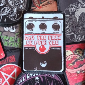Peut inclure: Un patch noir et blanc avec un graphique d'une pédale de guitare. La pédale a le texte "May the Fuzz Be With You" et les mots "The Fuzz" et "Awakens".
