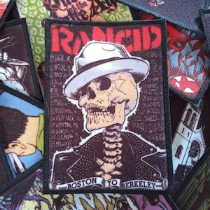 Puede incluir: Un parche bordado en blanco y negro con un esqueleto que lleva un fedora y una chaqueta de cuero. El texto "Rancid" está en letras rojas y blancas. El texto "Boston to Berkeley" está en la parte inferior del parche.