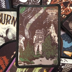 May include: A brown and green embroidered patch depicting a space scene. The patch features an astronaut, a spaceship, and a plant. The patch has a black border.