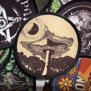 May include: A collection of embroidered patches featuring various designs, including a mushroom, a skull, and a star with a marijuana leaf. The patches are circular and have a black border.