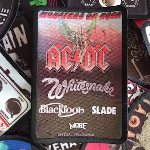 May include: A black and red patch with the text "Monsters of Rock" and the band name "AC/DC" in large white letters. The patch also features the names "Whitesnake", "Blackfoot", and "Slade" in smaller white letters. The text "More" and "Official Programme" are also visible on the patch.