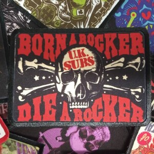 Può includere: Toppa nera e bianca con teschio e ossa incrociate e testo rosso che recita "Born a Rocker UK Subs Die a Rocker".