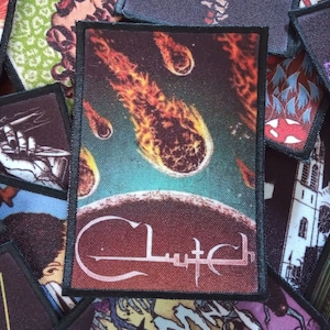 May include: A black and white woven patch featuring a red, orange and yellow image of a planet with flaming meteors falling from the sky. The text "Clutch" is written in a stylised font below the image.