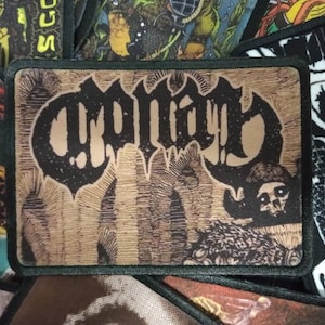 May include: A black and white embroidered patch with the word "CRYPTIC" in a gothic font. The patch has a brown background with a textured design and a skull image in the bottom right corner.