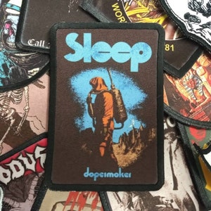 May include: A brown and blue embroidered patch with the word "Sleep" in a blue font. The patch features a person in a hooded jumpsuit walking through a desert landscape. The text "dope smoker" is printed at the bottom of the patch.