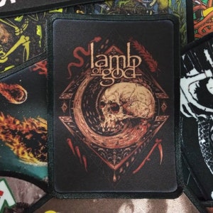 May include: A black and brown embroidered patch featuring the words "Lamb of God" and a skull with a snake wrapped around it.