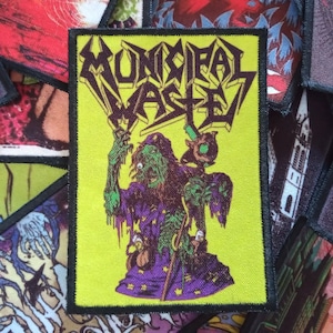 May include: A yellow patch with black stitching featuring a green and purple cartoon character holding a staff. The text "MUNICIPAL WASTE" is written in a black, stylized font.