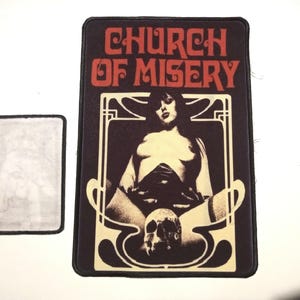 May include: A black and white patch with the text "Church of Misery" in red and white. The patch features a woman in a black and white outfit with a skull in her lap.