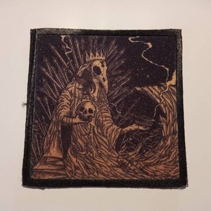 Può includere: Toppa ricamata in bianco e nero con una figura scheletrica che indossa una corona e una tunica, tenendo un teschio. La figura è circondata da uno sfondo scuro con un motivo a spirale.