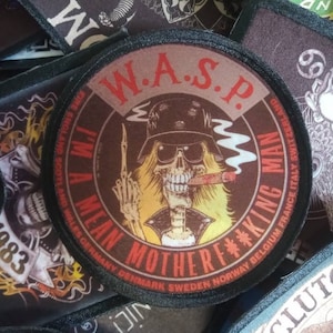 May include: A round, brown patch with the text "W.A.S.P. I'M A MEAN MOTHERF**KING MAN" in white and red lettering. The patch features a cartoon image of a skeleton with a cigarette in its mouth and a skull face wearing sunglasses and a black hat. The patch is surrounded by a black border.