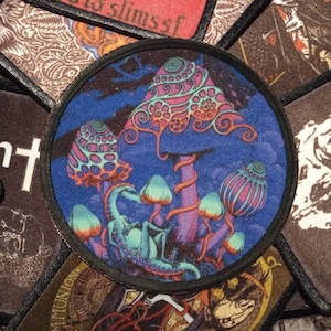 Può includere: Una toppa rotonda ricamata con un design psichedelico di funghi in tonalità di blu, viola, rosa e verde. I funghi hanno dettagli intricati e sono su uno sfondo blu scuro.