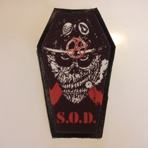 May include: A coffin-shaped patch with a black border. The patch features a skull graphic with a red anarchy symbol and the letters "S.O.D." in red. The background is dark burgundy.
