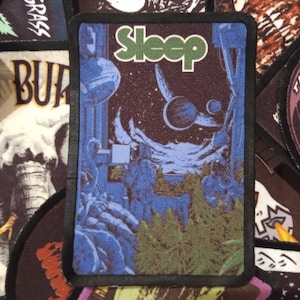 May include: A black and white embroidered patch with the word "Sleep" in green letters. The patch features a space scene with a planet, stars, and a spaceship.