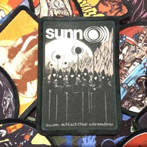 May include: A black patch with the band name "sunn" in white, and a graphic of a sun and figures. The text "02.09. schlachthof wiesbaden" is at the bottom. The patch is surrounded by other patches with various designs.