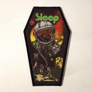 Può includere: Una toppa ricamata nera e verde a forma di bara con la parola "Sleep" in lettere verdi. La toppa presenta un astronauta dei cartoni animati con un casco bianco e una tuta verde e marrone. L'astronauta è in piedi davanti a uno sfondo rosso, arancione e giallo con una grande luna gialla.