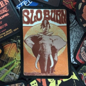 Op de afbeelding: Een vintage-stijl strijkbadge met een vrouw die op een olifant zit met de tekst "SLOBURN" in een retro-lettertype. De badge heeft een bruin, oranje en wit kleurenschema.