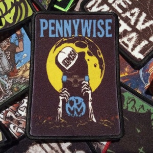 Könnte beinhalten: Ein schwarzer und gelber gestickter Patch mit dem Text "PENNYWISE" in blauen Buchstaben. Der Patch zeigt eine Skeletthand, die einen Kürbis mit einem blauen Gesicht und dem Wort "REMEMBER" darauf hält. Die Skeletthand befindet sich vor einem gelben Mond.