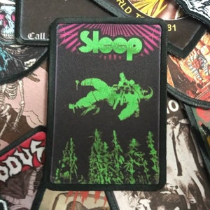 May include: Black and green embroidered patch with the band name "Sleep" in pink and green lettering. The patch features a green astronaut floating in space above a forest of green trees.