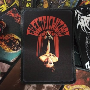 May include: Black and red embroidered patch with the text "Electric Wizard" in a white circle. The patch features a woman with long dark hair hanging upside down in a window.