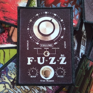 Può includere: Un cerotto bianco e nero con il testo "F-U-Z-Z" in lettere bianche. Il cerotto ha una grande manopola con un quadrante bianco e il testo "VOLUME" in lettere bianche. Il cerotto ha anche due manopole più piccole con il testo "BIAS" e "FUZZ" in lettere bianche. Il testo "True Bypass" è stampato in lettere bianche sotto le manopole.