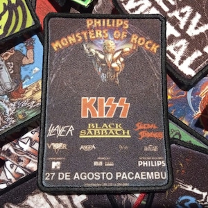 May include: A black and white patch with the text "PHILIPS MONSTERS OF ROCK" and the band KISS in red letters. The patch also features the bands Slayer, Black Sabbath, and Scorpions. The text "27 DE AGOSTO PACAEMBU" is at the bottom of the patch.