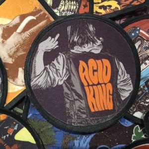 May include: A round, black and white patch with a person wearing a leather jacket with the words "Acid King" in orange letters on the back.