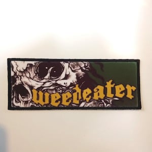 Könnte beinhalten: Ein schwarzer und grüner gestickter Patch mit einem Totenkopf und dem Wort "Weedeater" in gelben Buchstaben.