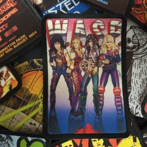 May include: A colorful iron-on patch featuring a band called "WASP" with four members in a rock and roll pose. The patch has a black border and a blue background with a gradient effect.