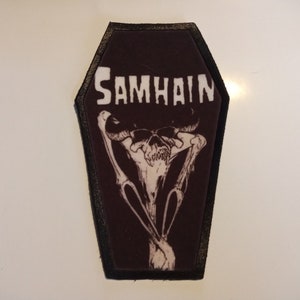 Könnte beinhalten: Ein Sargförmiger Aufnäher mit dem Wort "SAMHAIN" in weißen Blockbuchstaben. Darunter eine weiße Illustration eines Schädels mit Hörnern und gekreuzten Knochen auf dunklem Braun. Der Aufnäher hat einen schwarzen Rand.