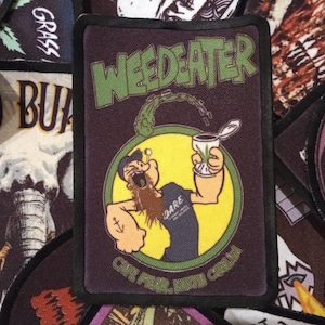 May include: A black and green embroidered patch with the text "WEED EATER" in green. The patch features a cartoon character with a beard holding a can with a marijuana leaf on it. The character is wearing a blue shirt with an anchor tattoo on his arm. The patch is surrounded by a yellow circle with a green border.