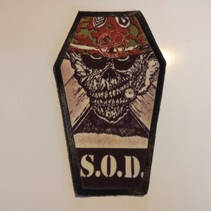 May include: A black and white embroidered patch with a skull wearing a military hat and a cigarette in its mouth. The patch is shaped like a coffin and has the text "S.O.D." in white letters on a black background.