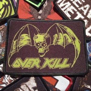 May include: A black and green embroidered patch featuring a skull with horns and wings. The text "OVER KILL" is written in green below the skull.