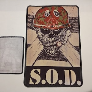 May include: A rectangular patch featuring a skull wearing a camouflage helmet with a peace symbol. The skull is smoking a cigarette. The text "S.O.D." is at the bottom. A smaller, blank patch is next to it.