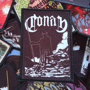 May include: A black and white embroidered patch with the word "Conan" in a gothic font. The patch features a silhouette of a figure with horns standing on a rocky cliff.
