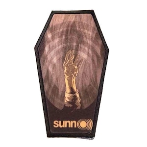 May include: A brown coffin-shaped patch with a hand reaching out from the inside. The hand is a light brown color and the background is a dark brown with a light brown pattern. The text "sunno" is printed in yellow at the bottom of the patch.