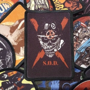 Peut inclure: Un écusson brodé noir et orange avec un crâne portant un casque militaire et un éclair en forme de croix. Le texte "S.O.D." est sous le crâne.