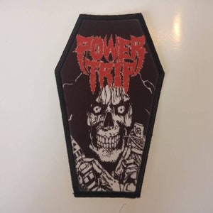 May include: A black coffin-shaped patch featuring the band name "POWER TRIP" in red, stylized lettering. The patch has a graphic of a skeletal figure with money in its hands. The patch has a black border.
