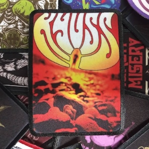 Puede incluir: Un parche bordado en negro y rojo con la palabra "RUSS" en una fuente estilizada. El fondo del parche es de color naranja y rojo fuego con una imagen borrosa de una cara.