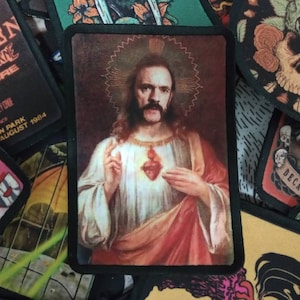 May include: A black and white embroidered patch featuring a man with a long beard and moustache, wearing a red robe and holding a heart in his hand. The man is depicted in a religious style, with a halo above his head. The patch is surrounded by a black border.