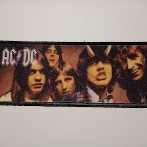 Puede incluir: Parche rectangular con borde negro, con la banda AC/DC. El parche muestra una imagen en tonos sepia de los miembros de la banda. El logotipo de la banda, "AC/DC" en blanco con un rayo, está en la parte superior izquierda.