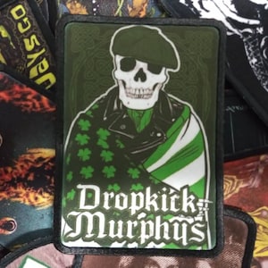 Op de afbeelding: Een zwart en groen geborduurd patch met een schedel die een groene hoed en een groen-witte sjaal draagt. De tekst "Dropkick Murphys" staat in witte letters op de patch.