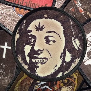 Peut inclure: Un patch brodé en noir et blanc représentant un portrait rapproché d'une personne avec une feuille de marijuana sur le front et le mot "feeb" sur les dents.