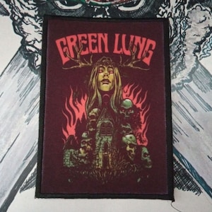 May include: A rectangular patch with a dark red background features the text "GREEN LUNG" in red, stylized lettering. The image depicts a woman with antlers, surrounded by flames and skulls. The patch has a black border.