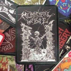 May include: Black and white embroidered patch with the text "MONOCHIAL WASTE" in a gothic font. The patch features a skeletal figure surrounded by barbed wire and a dark background.