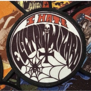 May include: A black and white round patch with the text "I Hate Electric Wizard" in a stylised font. The patch features a spider's web with a spider in the centre and a skull with a cross behind it.