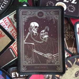 May include: A black and white tarot card with a dark red background. The card depicts two skeletons holding hands. The card is labeled "The Devil" and has the Roman numeral "XV" in the top right corner.