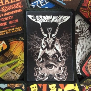 May include: Black and white embroidered patch featuring a winged demon figure, an eye, and the letters "W", "A", and "Ω".